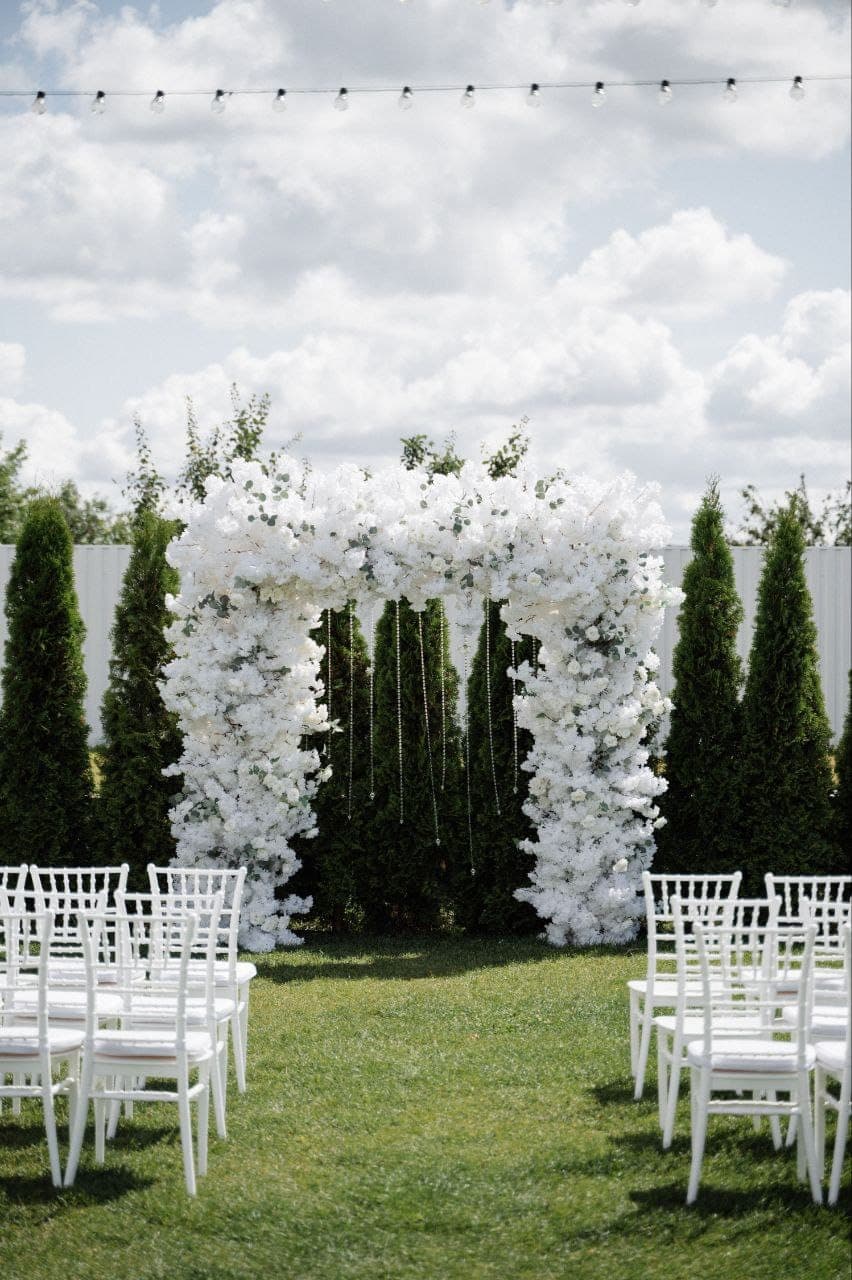 White floral ceremony arch flanked by conifers, with chiavari chairs on the lawn