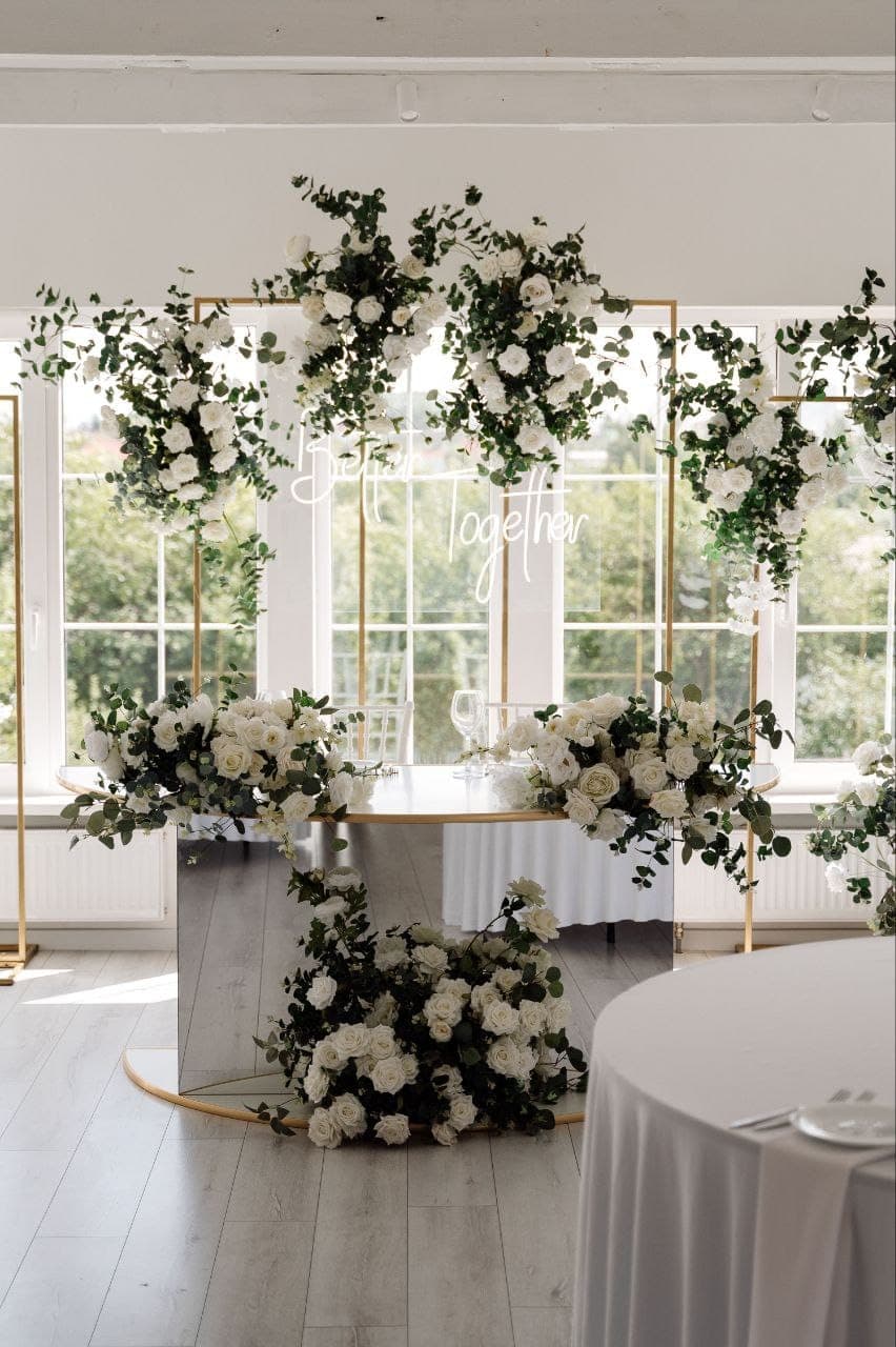 White and green floral installation on gold stands beside tall windows
